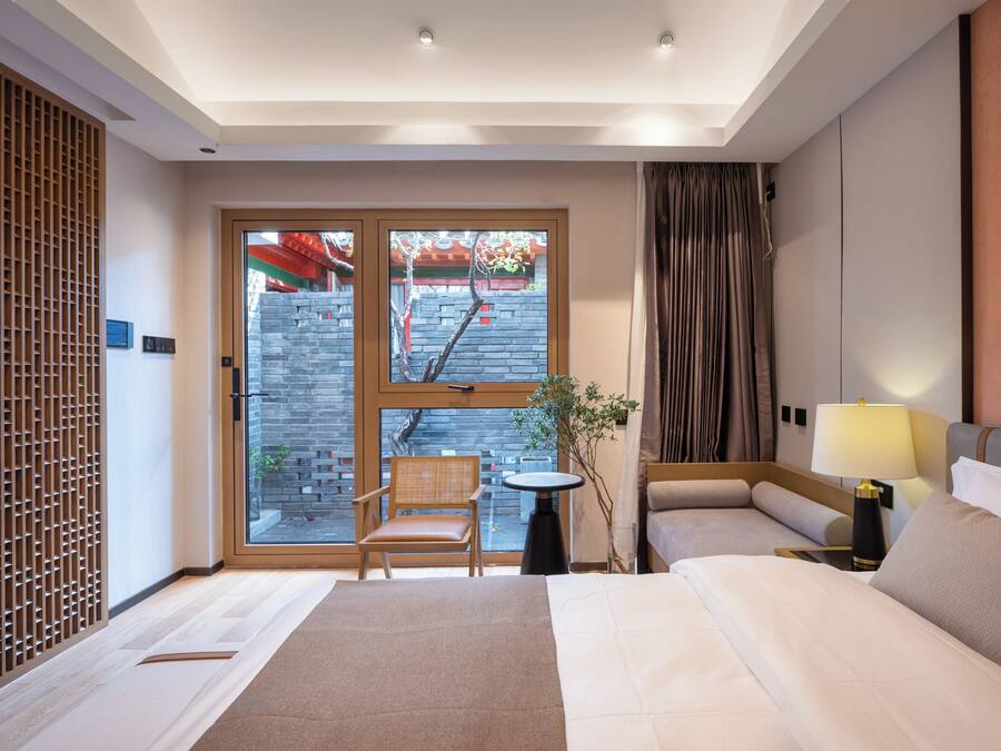 Ein Hotelzimmer mit einem Bett, zwei Nachttischen, einem Sessel, einem Fenster mit Blick auf eine Ziegelwand und Bäume sowie einer Wandverkleidung.