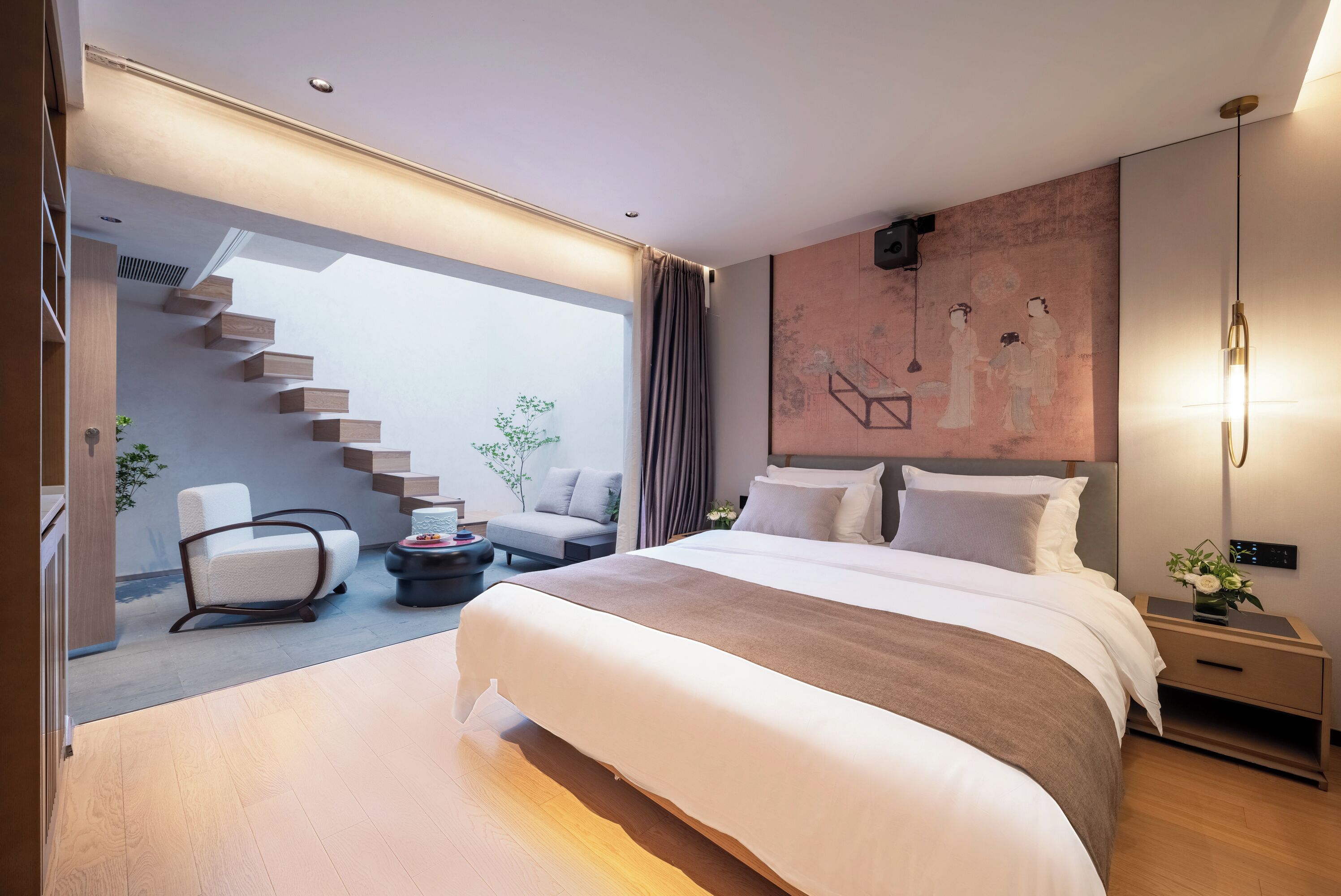 Una habitación de hotel moderna con una cama grande, una zona de estar con sofá y sillón, una escalera que lleva a un nivel superior y una obra de arte colgada en la pared.