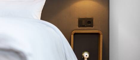 A neatly made bed with white linens, a wall-mounted nightstand with a small figurine, and a black lamp.