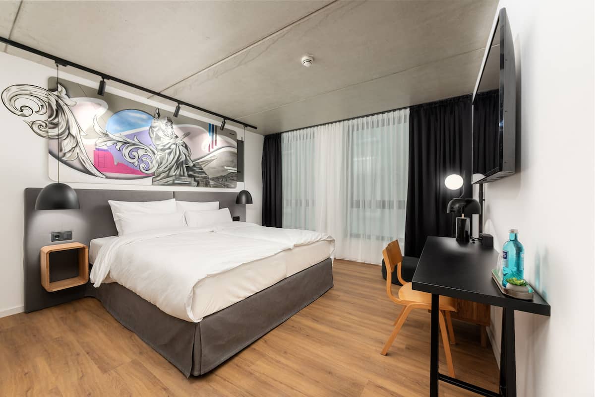 Une chambre d’hôtel moderne équipée d’un lit, d’un bureau, d’une chaise, d’un téléviseur à écran plat et d’un élément décoratif mural.