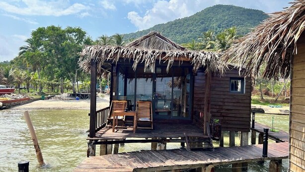 A wooden structure with a thatched roof, a glass door, and a small table with chairs on a dock extending over water. - Freedom Beach Resort (Phu Quoc)