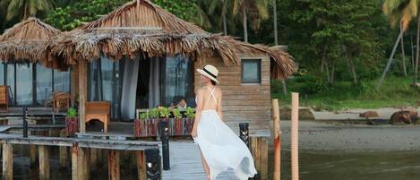 A wooden dock leading to a thatched-roof structure with a view of palm trees and water.