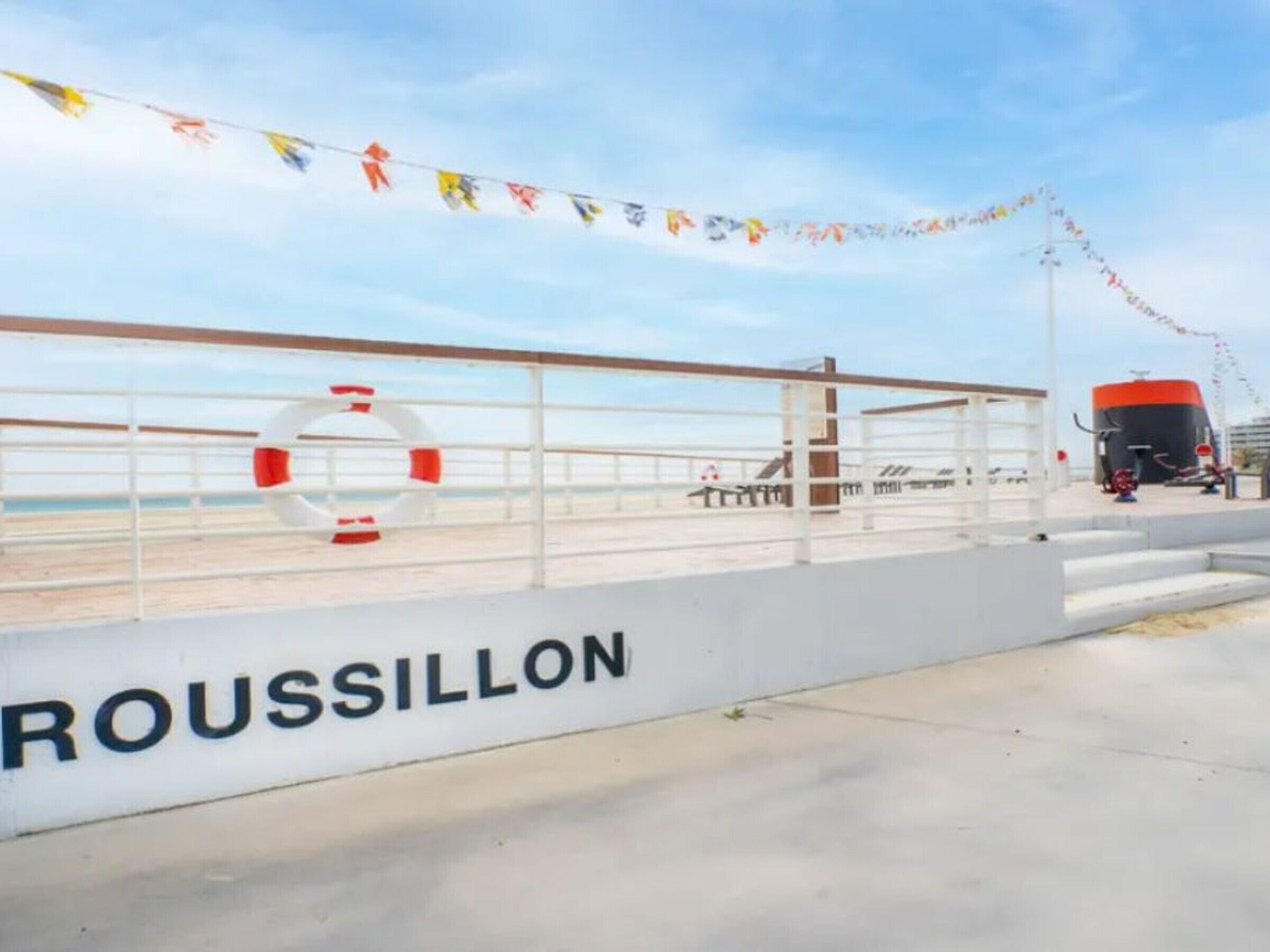A waterfront promenade with a railing, a lifebuoy, and a sign that reads "ROUSSILLON".