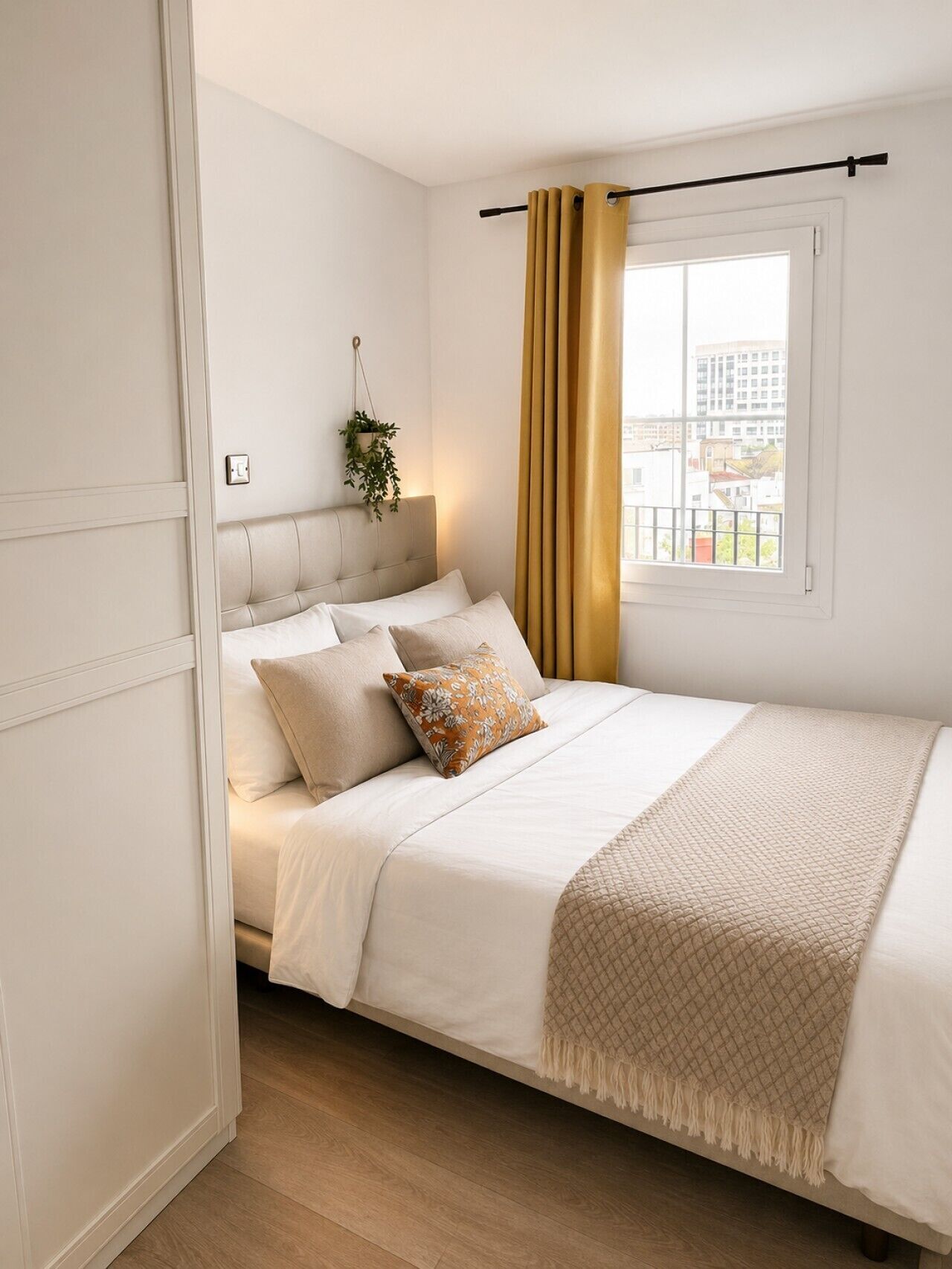 Ein Schlafzimmer mit einem Bett, einem Vorhangfenster, einer Wandpflanze und Blick auf Gebäude.