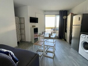 A compact living space with a sofa, a dining table with chairs, a TV mounted on the wall, a washing machine, and a sliding glass door leading to an outdoor area.
