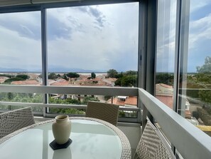 Ein Balkon mit Korbmöbeln und einer Vase, mit Blick auf eine Stadt mit roten Dächern und einem Gewässer.