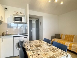 Une cuisine compacte équipée d’un four à micro-ondes, d’un lave-linge et d’un évier. On y trouve une table à manger recouverte d’une nappe à motifs et entourée de chaises bleues. Un canapé avec des coussins à rayures se trouve dans l’espace salon.