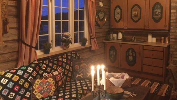A cozy cabin interior with a large window, a quilted sofa, a wooden cabinet with mirrors, and a candlelit table. - The Reverse Escape Room Cabin (Gausdal)