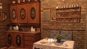 A rustic dining area with a wooden table set for two, surrounded by wicker chairs. A cabinet with decorative doors is to the left, and a shelf with ceramic figurines is above the table. - The Reverse Escape Room Cabin (Gausdal)