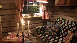 A cozy cabin interior with a wooden log wall, a window with curtains, a table with candles, a lamp, a quilted sofa, and a basket of yarn. - The Reverse Escape Room Cabin (Gausdal)