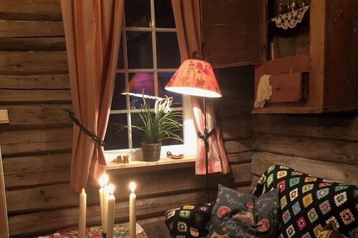 A cozy cabin interior with a wooden log wall, a window with curtains, a table with candles, a lamp, a quilted sofa, and a basket of yarn.
