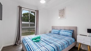 Une chambre à coucher avec un lit, une table de chevet, une lampe, une fenêtre avec des rideaux et un tableau encadré au mur.