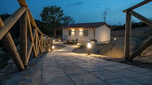 Un sendero lleva a una casa con tejado de tejas y dos ventanas iluminadas, rodeada por una valla de madera y macetas con plantas.