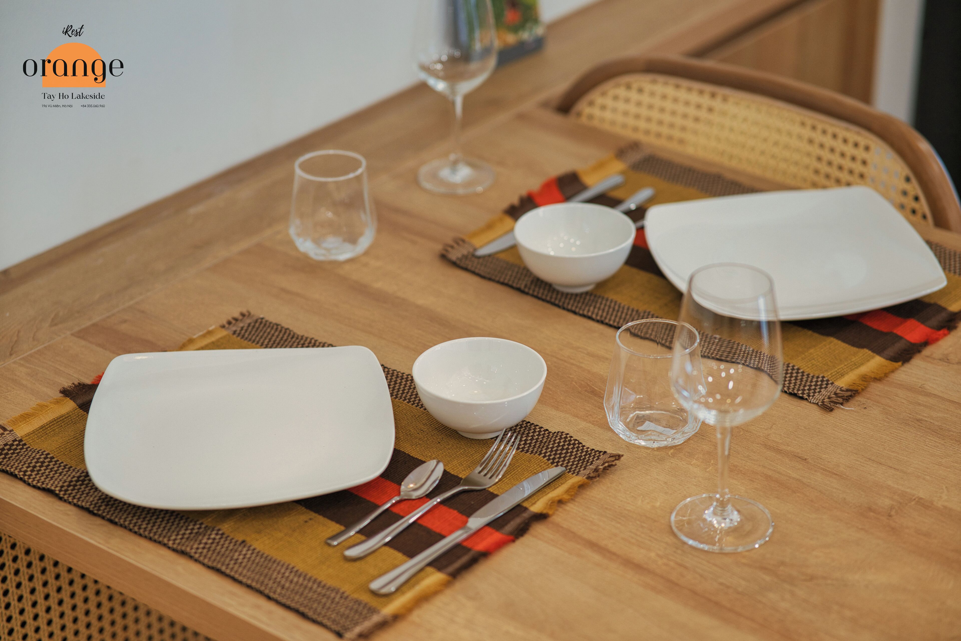 A dining table set with white plates, bowls, cutlery, and wine glasses, with a placemat and a woven chair backrest.