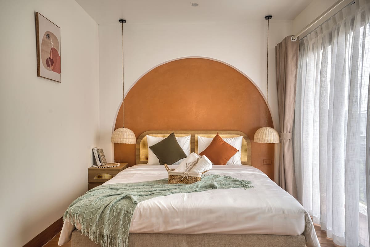 Een netjes opgemaakt bed met hoofdbord, een nachtkastje met een lamp, en een raam met gordijnen.