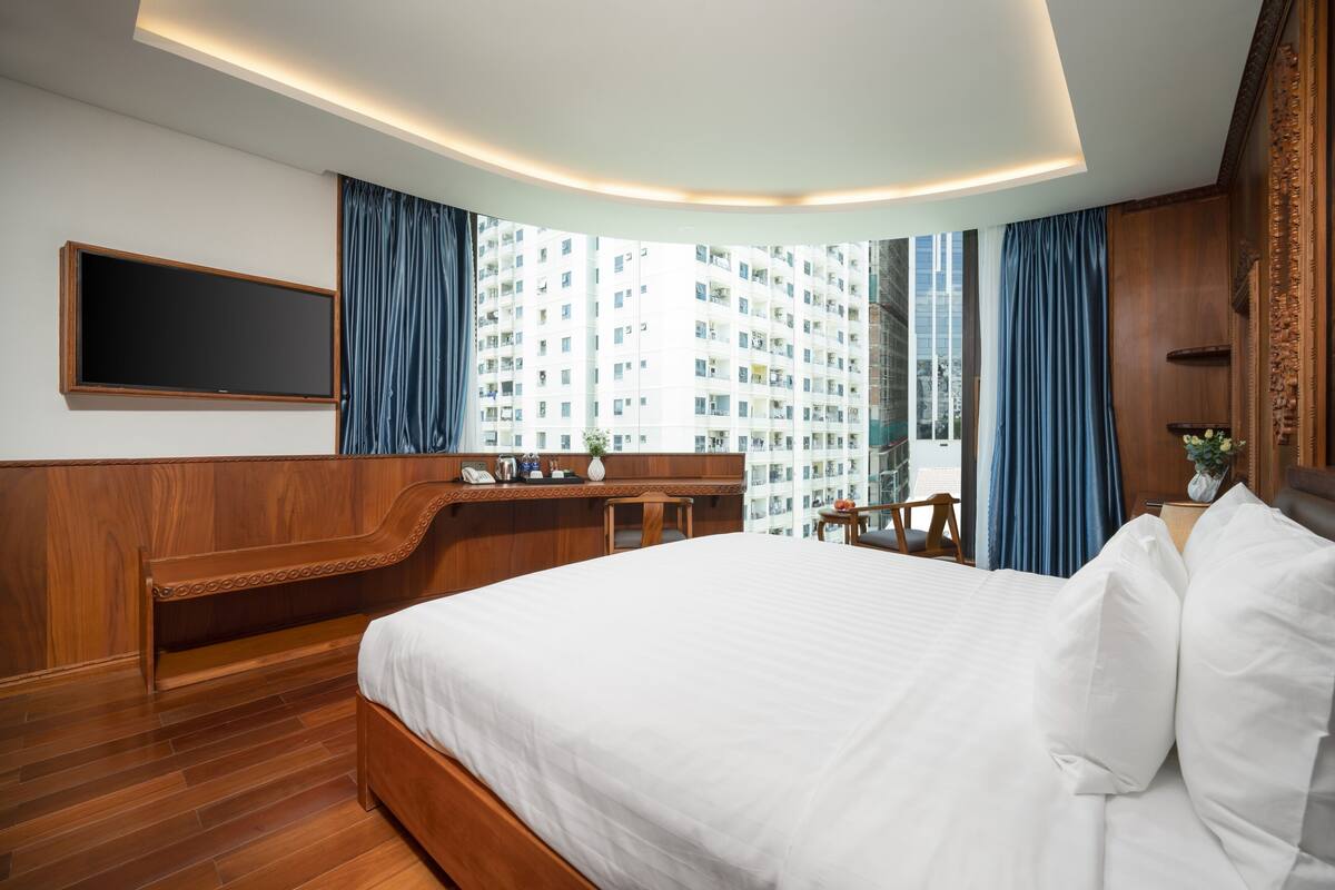 A modern hotel room with a large bed, wooden bench, desk, and a large window offering a view of a high-rise building.