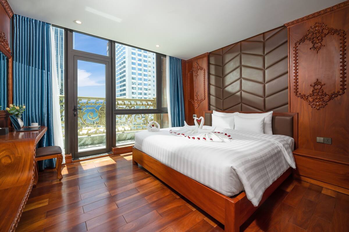 A spacious bedroom with a large bed, wooden flooring, a desk, and a balcony with a view of a high-rise building.