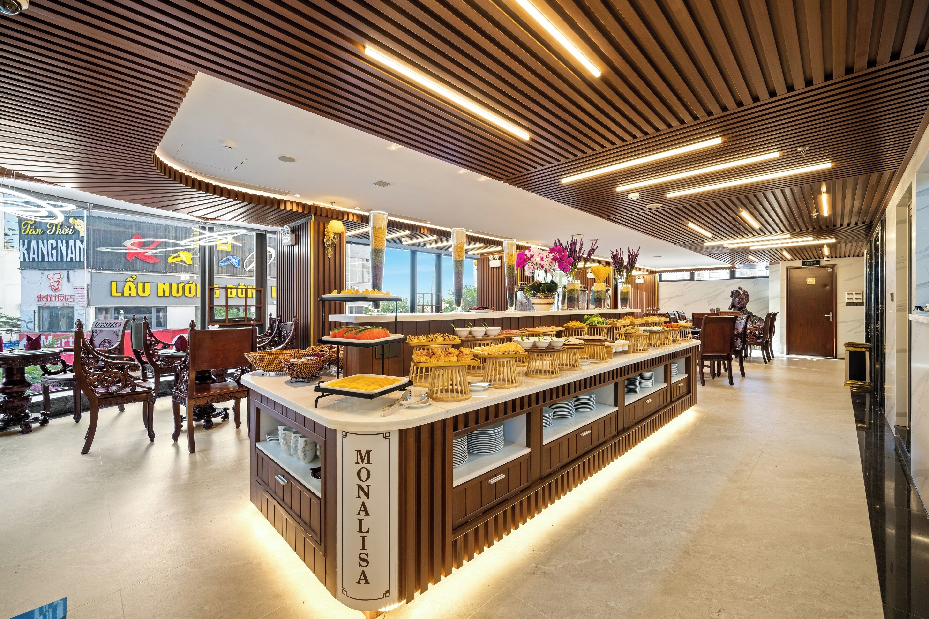 A modern buffet area with a wooden counter labeled "MONALISA," featuring various food items and a seating area with chairs.