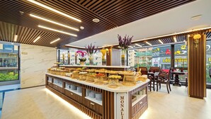 A modern cafe with a wooden counter displaying various food items, a seating area with tables and chairs, and a view of the outside through large windows. - Monalisa Luxury Hotel (Da Nang)