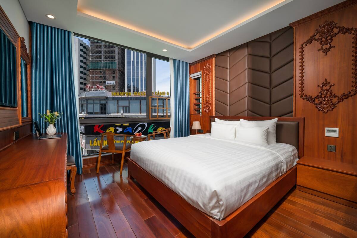 A modern hotel room with a large bed, wooden flooring, a desk with a chair, and a view of the city through large windows.
