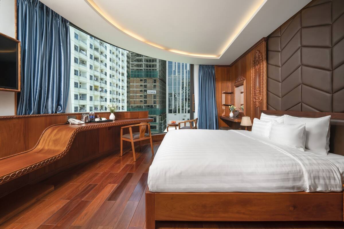 A hotel room with a large bed, wooden furniture, a desk, and a view of high-rise buildings.