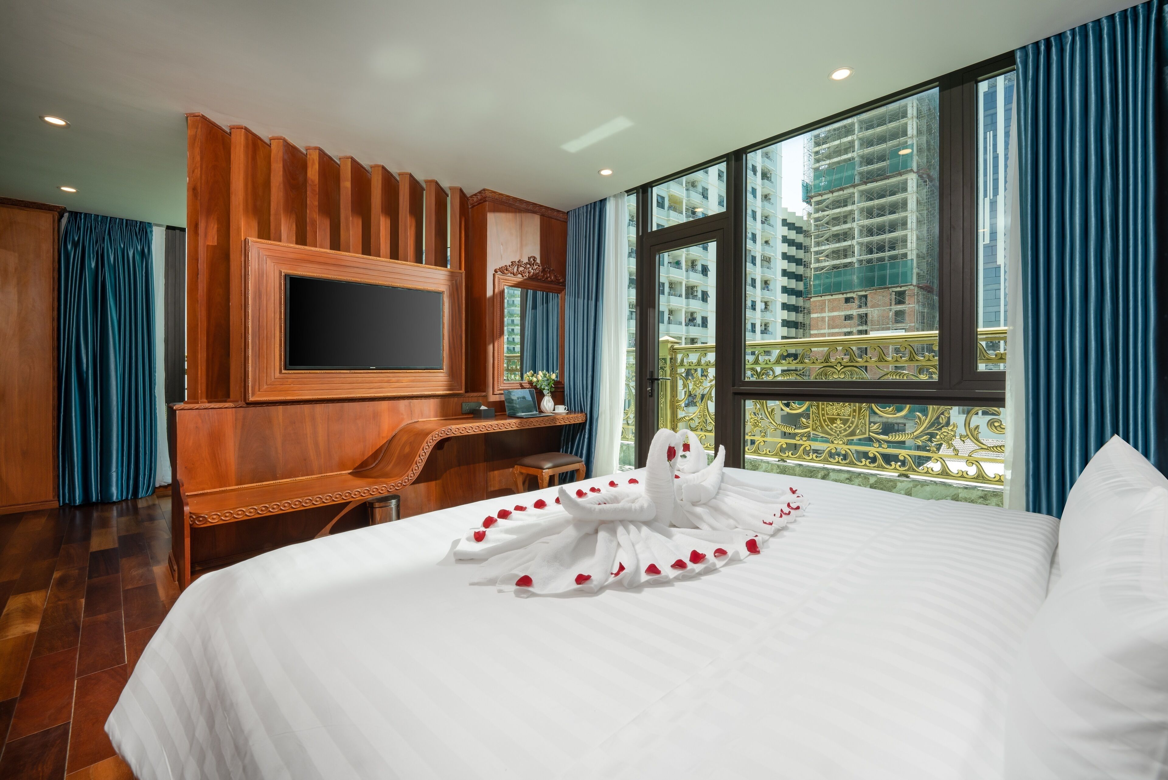 A hotel room with a large bed, a wooden desk, a flat-screen TV, and a balcony with a view of high-rise buildings.