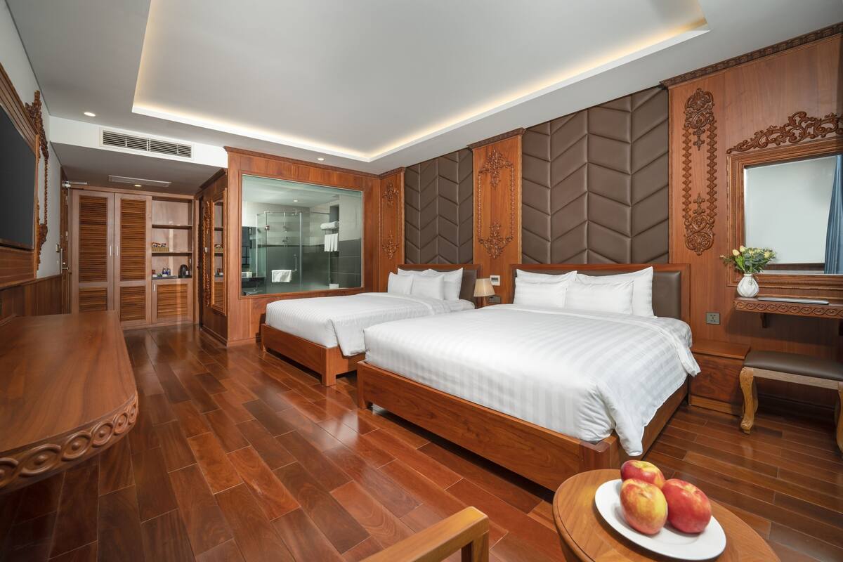 A hotel room with two beds, a wooden dining table, a mirror, and a bathroom visible through a glass door.