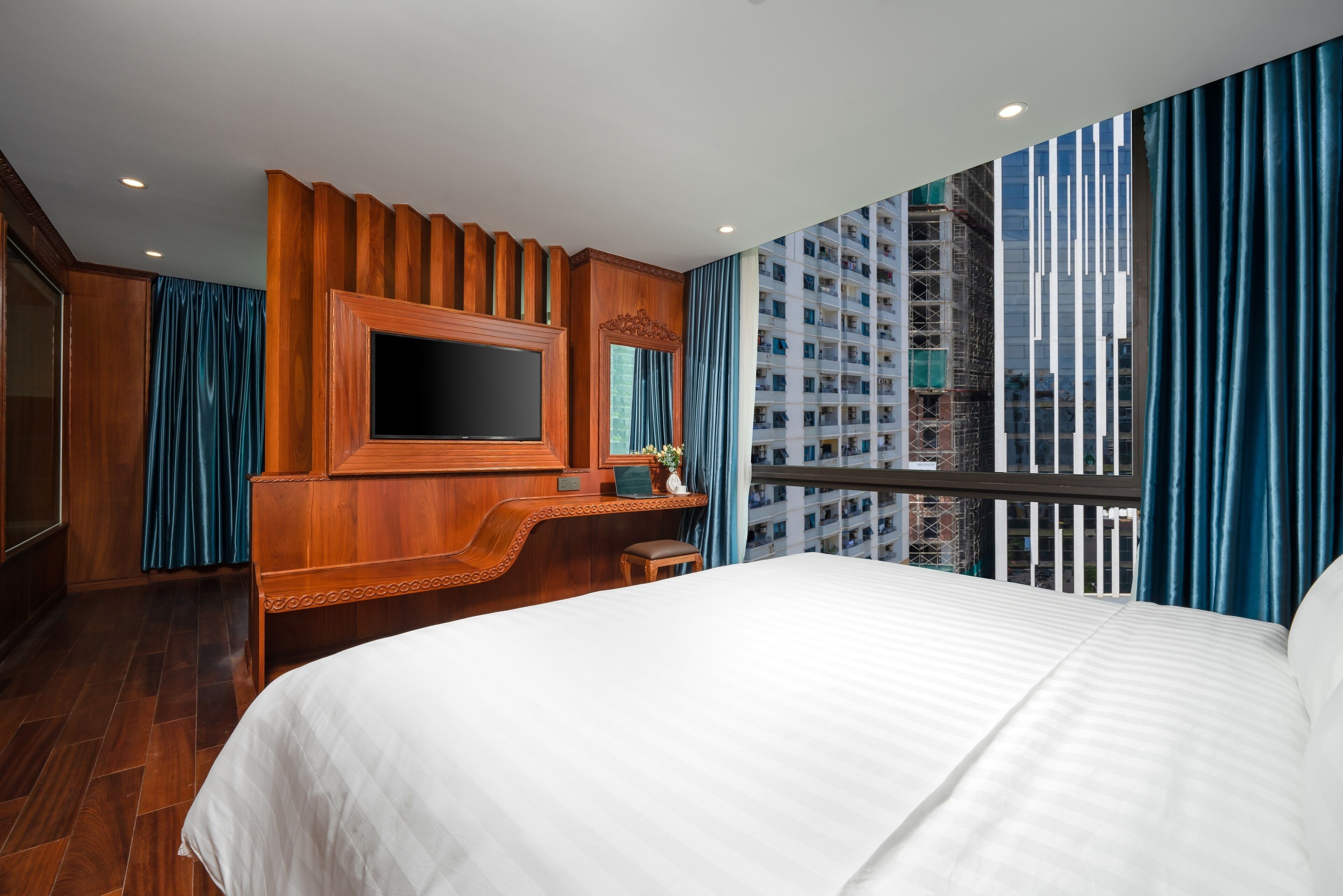 Une chambre d’hôtel avec un grand lit, un bureau en bois équipé d’un téléviseur à écran plat, un miroir et une vue sur la ville, accessible par un balcon agrémenté de rideaux bleus.