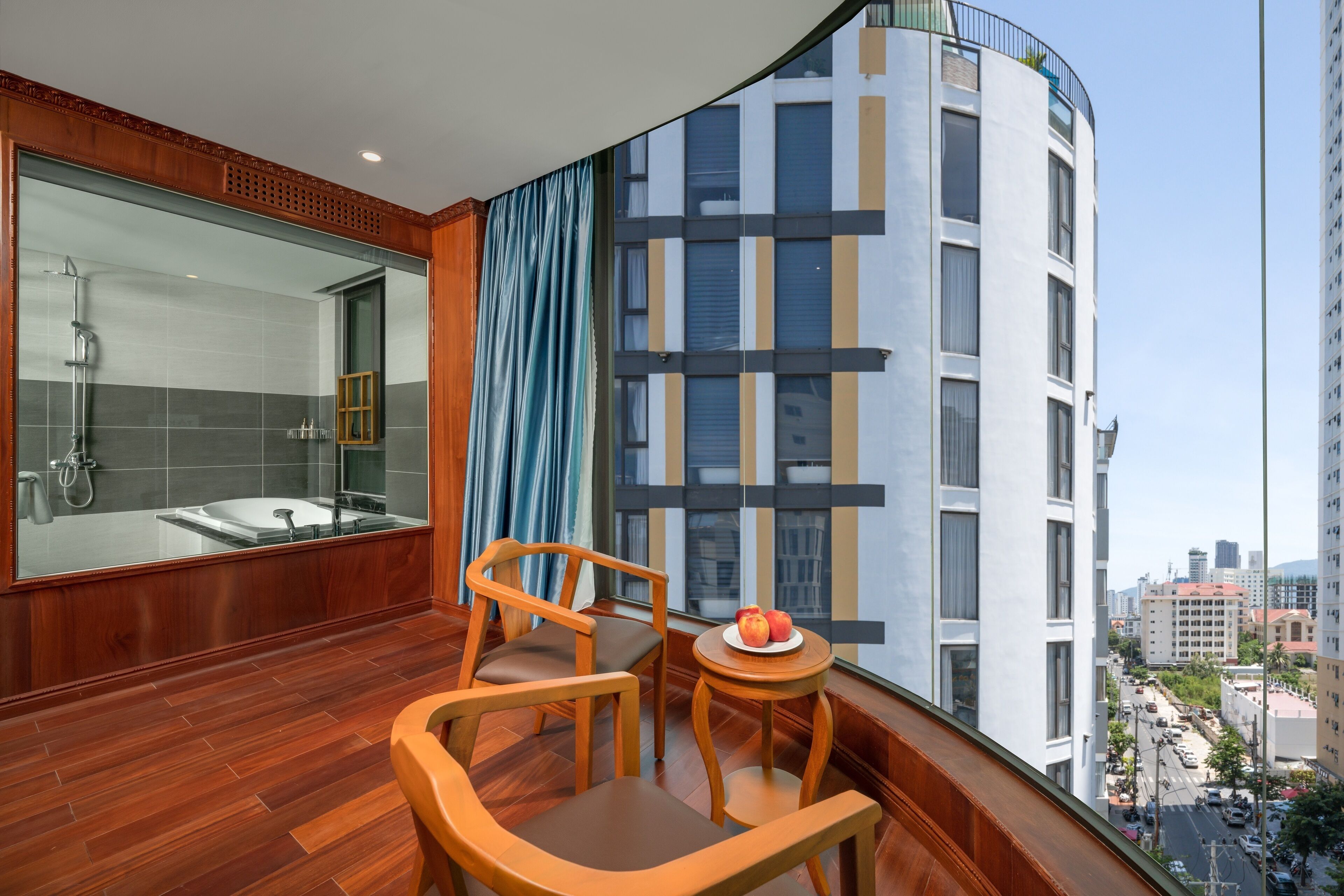 A balcony with a wooden floor, two wooden chairs, a small round table with a plate of fruit, and a large window offering a city view.