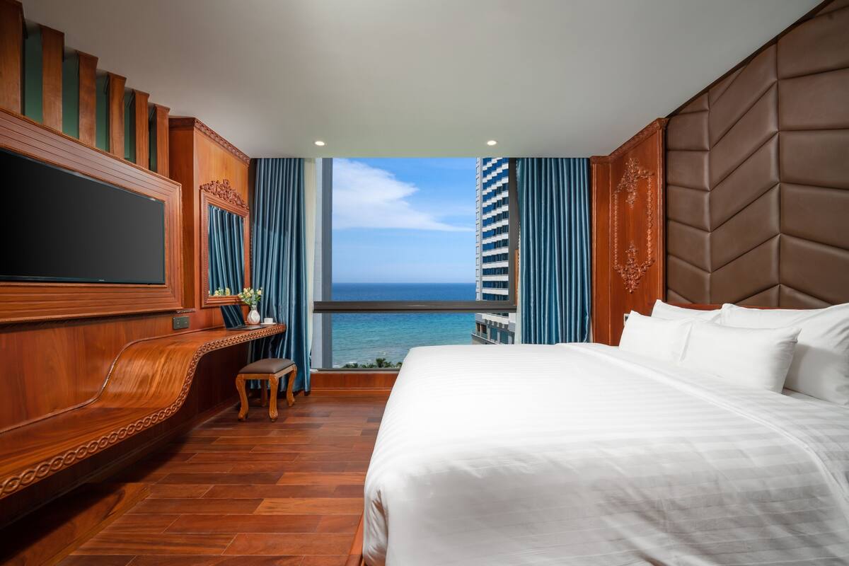 A hotel room with a large bed, a wooden bench, a flat-screen TV, and a view of the ocean.