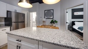 A modern kitchen with granite countertops, stainless steel appliances, pendant lighting, and a view into a bedroom.
