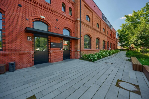 A red brick building with large arched windows, a paved walkway, and a green area with benches.