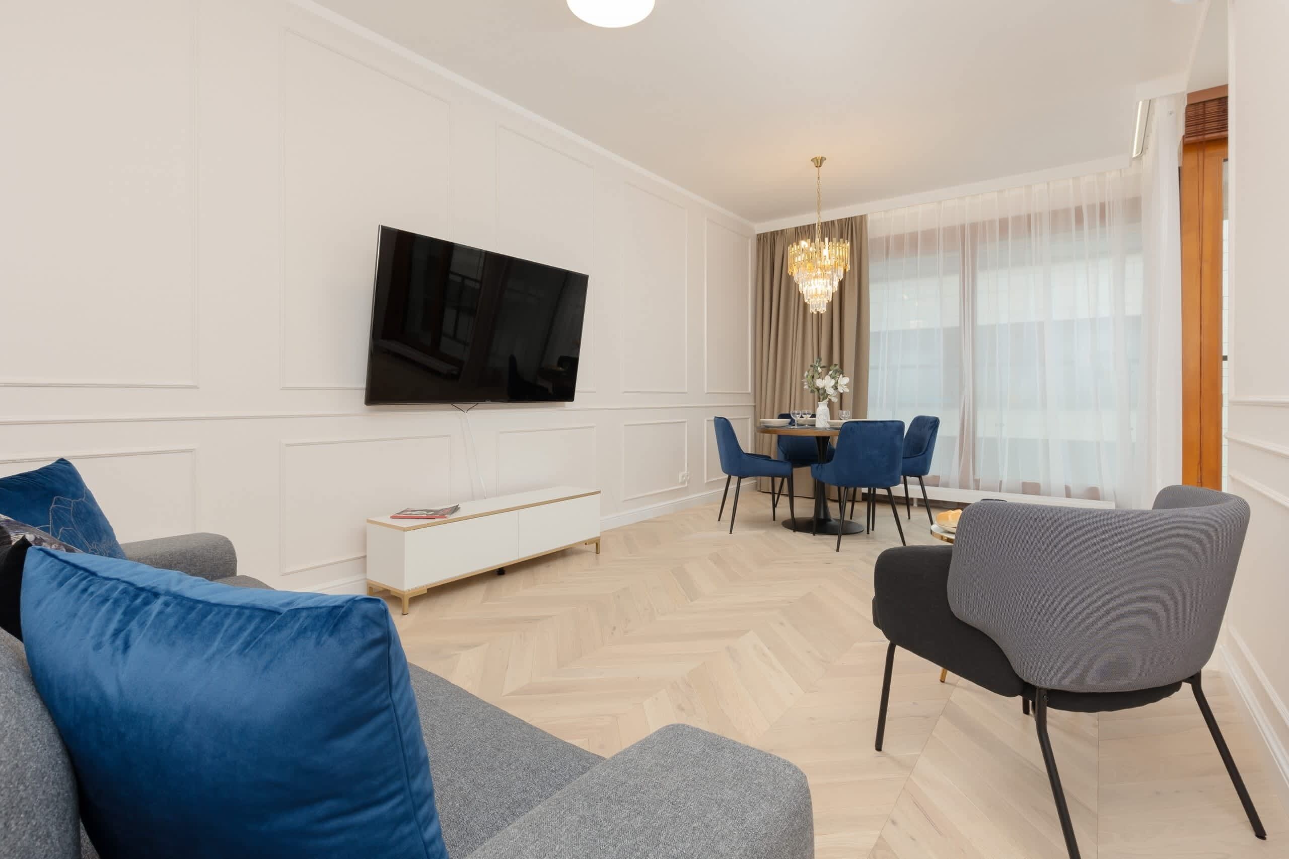 A modern living room with a flat-screen TV mounted on the wall, a dining area with blue chairs, and a chandelier.