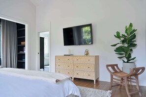 Une chambre à coucher avec un lit, un téléviseur fixé au mur, une commode en bois, une plante en pot et deux chaises en bois.