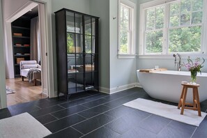 A modern bathroom with a freestanding bathtub, a glass cabinet, and a large window with a view of greenery.
