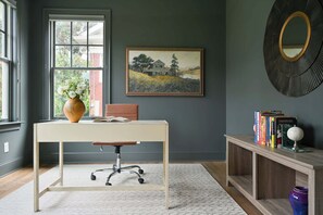 Una habitación con escritorio, silla, jarra, estantería y un cuadro en la pared.