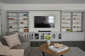 A modern living room with a grey sofa, a coffee table with books, a flat-screen TV mounted on the wall, and built-in shelving with various items.