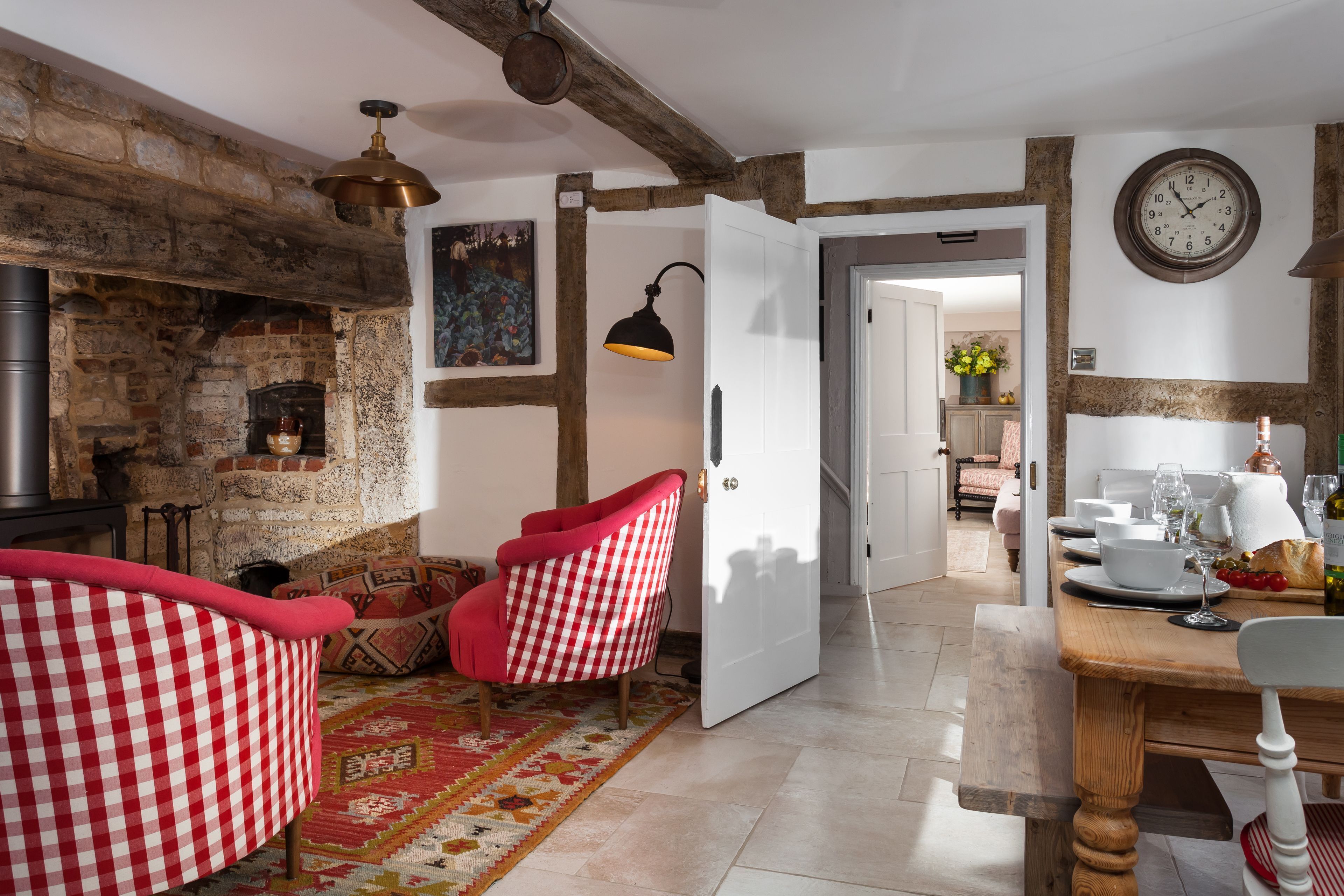 A cozy room with exposed brick walls, a fireplace, a checkered armchair, a dining table with chairs, and a clock on the wall.