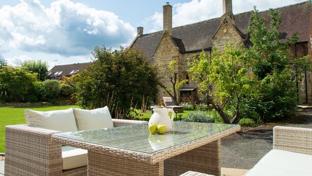 Un espace extérieur aménagé avec des meubles en osier et une table en verre, situé devant un bâtiment en pierre, avec un jardin en arrière-plan.