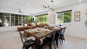 A spacious living room with a large window, a dining area with a wooden table set for four, and a brown leather sofa. - Family-Friendly Gainesville Home - Walk to UF - Pool + Sleeps 16 (Gainesville)