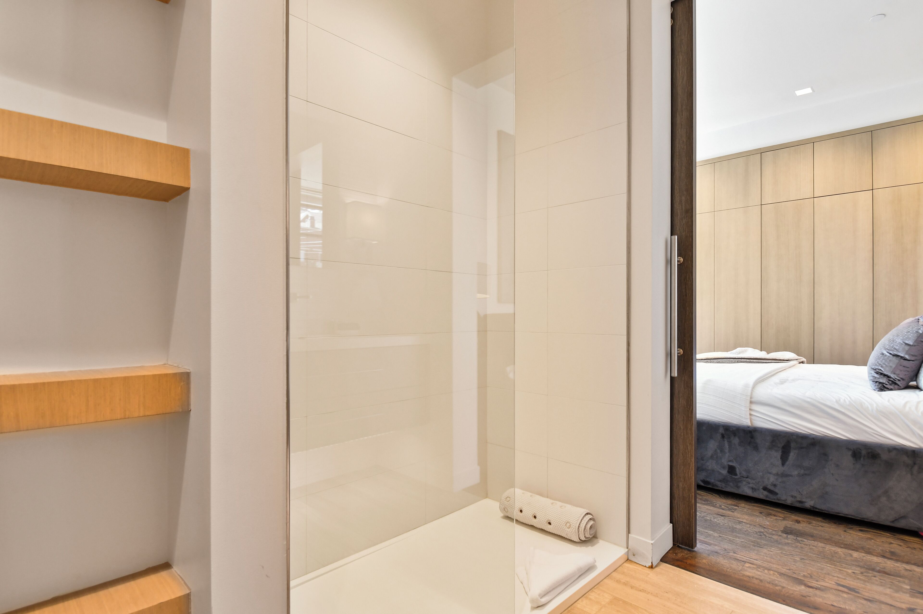 Une salle de bain moderne avec une douche à parois de verre, des étagères en bois et un lit visible dans la pièce adjacente, avec un matelas et des oreillers gris.
