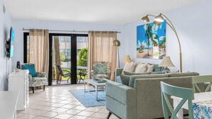 Ein Wohnzimmer mit einer Couch, Sesseln, einem Couchtisch und einer Lampe. Ein Schiebeglasfenster führt in einen Außenbereich.