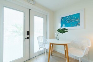 A room with a white door, a white table, two white chairs, a potted plant, and a framed picture on the wall.