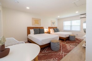 A hotel room with two beds, a small table, a potted plant, and a window with blinds. - THE JUNI - Studio Steps to Beach, Downtown Cocoa (Cocoa Beach)