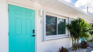 A turquoise door, a window with blinds, and a small garden with plants.