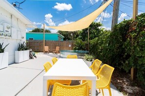 Une terrasse extérieure avec une table blanche et des chaises jaunes, une clôture en bois et un bâtiment vert en arrière-plan.