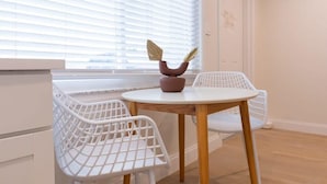 Una habitación con una mesa redonda de madera, dos sillas de alambre blancas, un florero con hojas secas y una ventana con persianas.