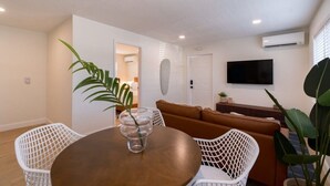 A dining area with a round table, white chairs, a brown sofa, and a TV mounted on the wall. - Welcome to The Juni-The Streets (Cocoa Beach)