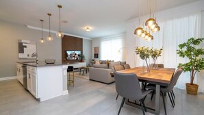 Eine moderne Küche mit weißen Schränken, ein Essbereich mit Holztisch und Stühlen sowie ein Wohnzimmer mit Sofa und Fernseher.