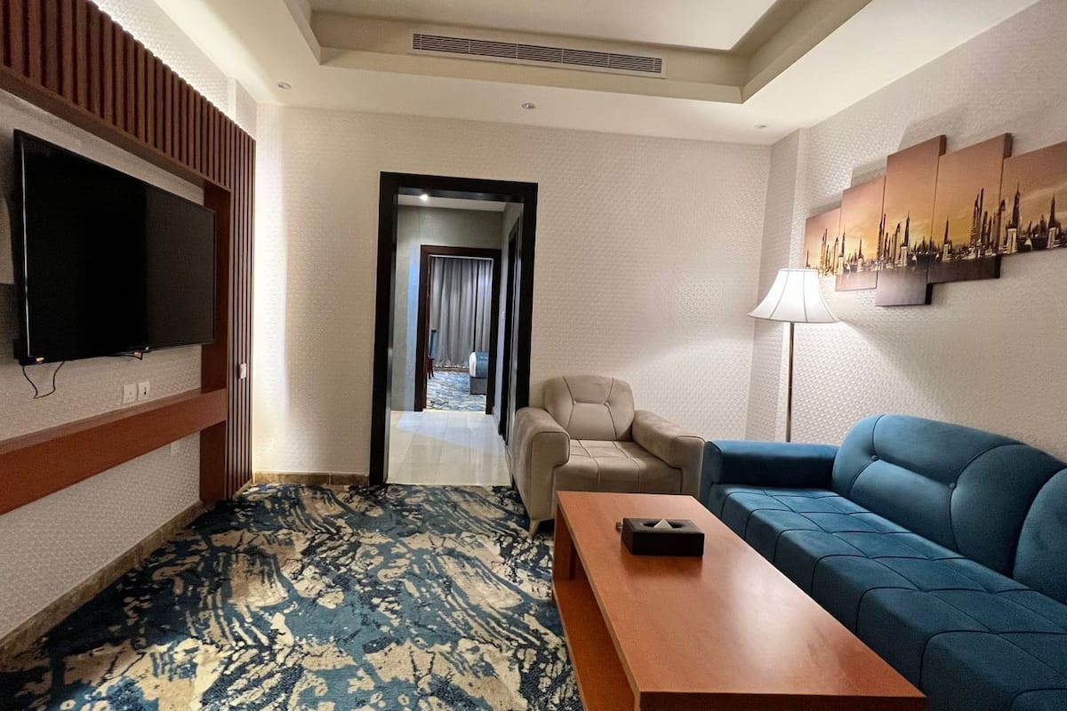 Une chambre d’hôtel équipée d’un téléviseur à écran plat fixé au mur, d’un canapé bleu, d’un fauteuil beige et d’une table basse en bois.